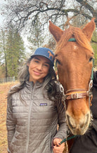 Pretty woman with long dark hair stands next to a chestnut horse and she wears a cute trucker hat that is dark blue on the front panel with white mesh on the back and a snapback closure. A chestnut horse is printed on the front side of the hat.