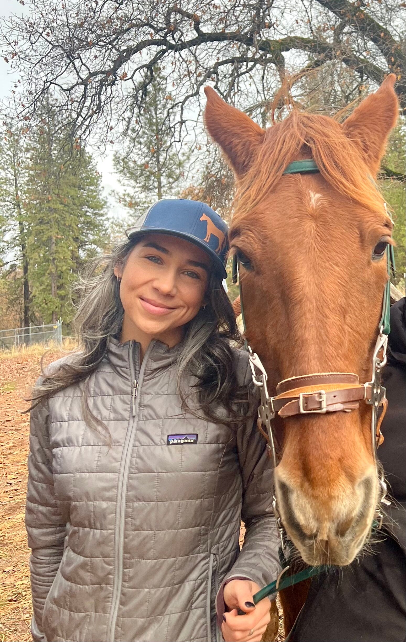 Pretty woman with long dark hair stands next to a chestnut horse and she wears a cute trucker hat that is dark blue on the front panel with white mesh on the back and a snapback closure. A chestnut horse is printed on the front side of the hat.