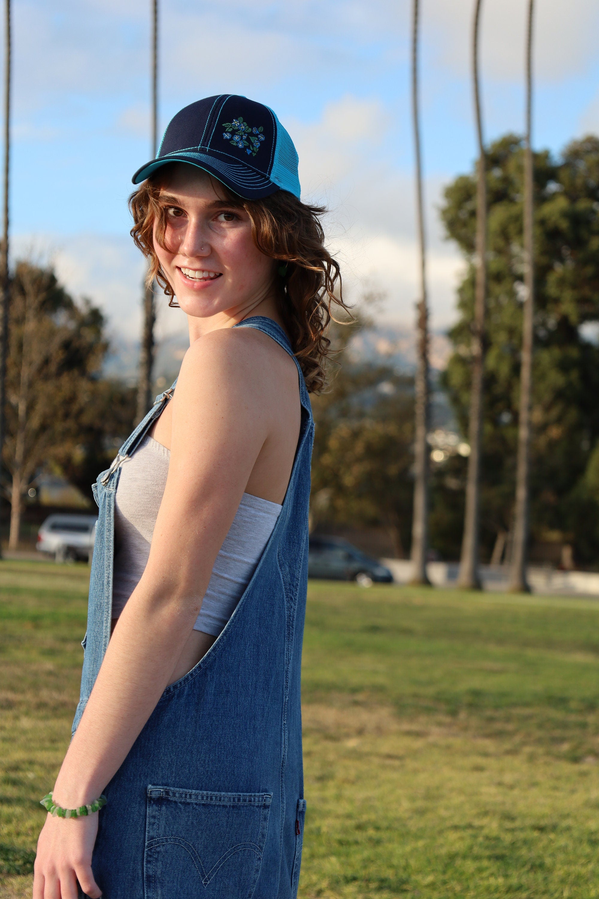 Girl with brown curly hair wears a cute trucker hat that is navy blue on the front with light blue mesh on the back and a velcro closure. Morning glory flowers are embroidered on the front side of the hat.