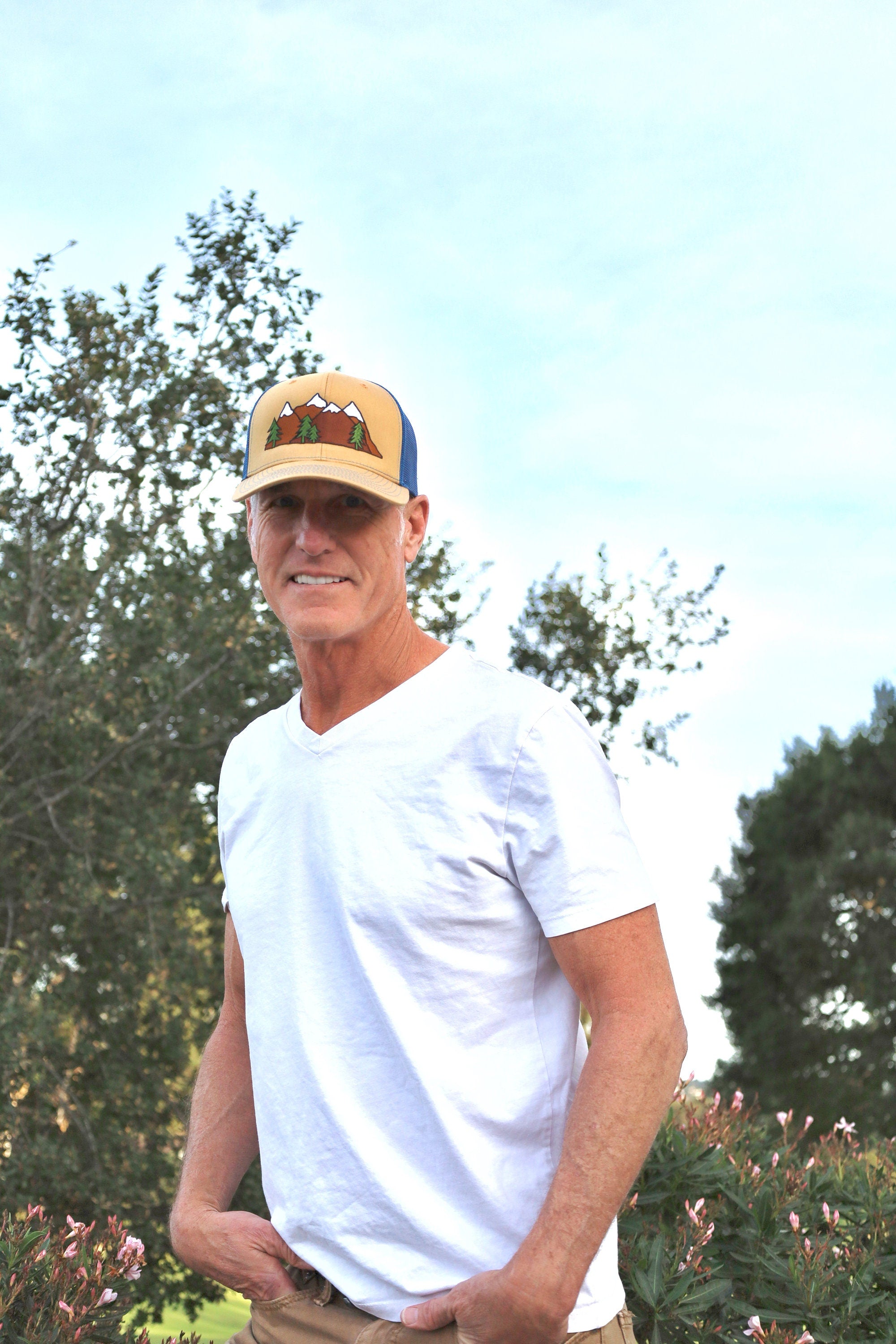 Outdoorsy man wears a vintage inspired trucker hat that is khaki on the front panel with blue mesh on the back and a snapback closure. A mountain scene is embroidered on the front of the hat.