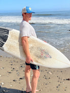 Young man is on the beach with a surf board looking at the waves. He wears a trucker hat that is blue on the front with tan mesh on the back and a snapback closure. A circle embroidered patch with ocean and sun is on the center front of the hat.