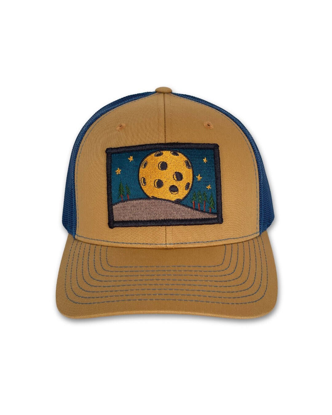 Trucker hat with a low profile. Front bill and panel is tan cotton fabric. A yellow pickleball moon over a tan hill with green trees is embroidered on a patch in the center. The back panel is blue made of mesh with a snapback closure.